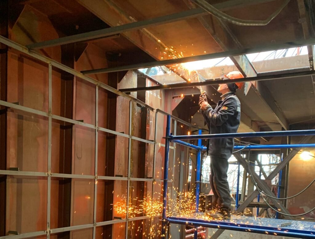 A construction worker welds metal indoors, creating flying sparks in a factory setting.